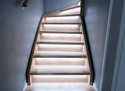 Moderní dřevěné schodiště s LED světly pod každým stupněm, které podtrhují minimalistický design s teplým osvětlením na pozadí šedých stěn.