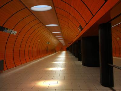 Jasná LED stropní svítidla osvětlují moderní tunel metra, jehož stěny jsou oranžové a podlaha obložená dlaždicemi.