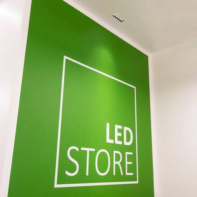 Alternativní text: "Zelená stěna s bílým čtvercovým obrysem "LED STORE", tučná písmena, stropní světla"