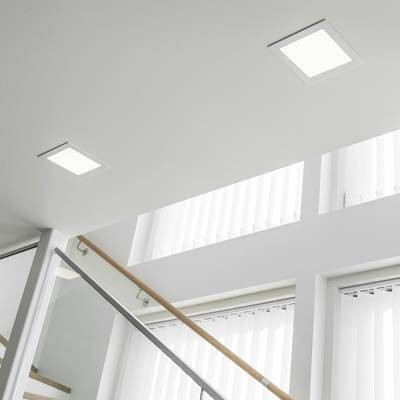 Jasná čtvercová vestavná stropní svítidla osvětlují moderní schodiště se skleněným zábradlím a velkými okny se svislými závěsy.