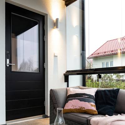 Moderní sluneční pokoj s energeticky úsporným LED osvětlením, černými dveřmi, šedou pohovkou s barevnými polštáři, prosklenými stěnami, které ukazují dům se zářivě červenou střechou.