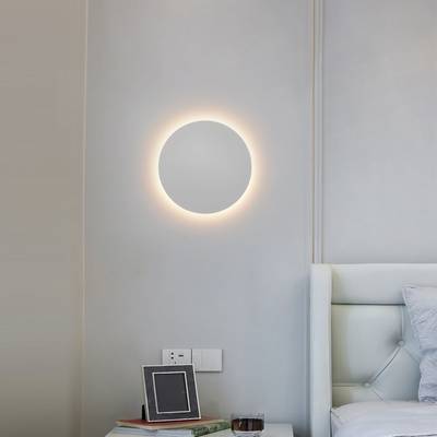 Moderní kruhové nástěnné svítidlo osvětluje minimalistickou ložnici se světle šedými stěnami, bílým čalouněným čelem postele a stylovou dekorací nočního stolku. Obchod s LED osvětlením.