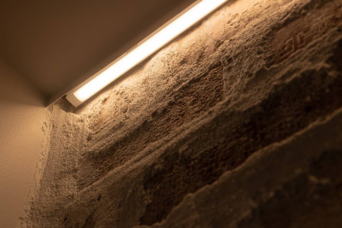 Detailní záběr instalace LED pásku na dekorativní stěně. LED pásek je nainstalován do hliníkového profilu pro povrchovou montáž, který je umístěn mezi lištou a stěnou.