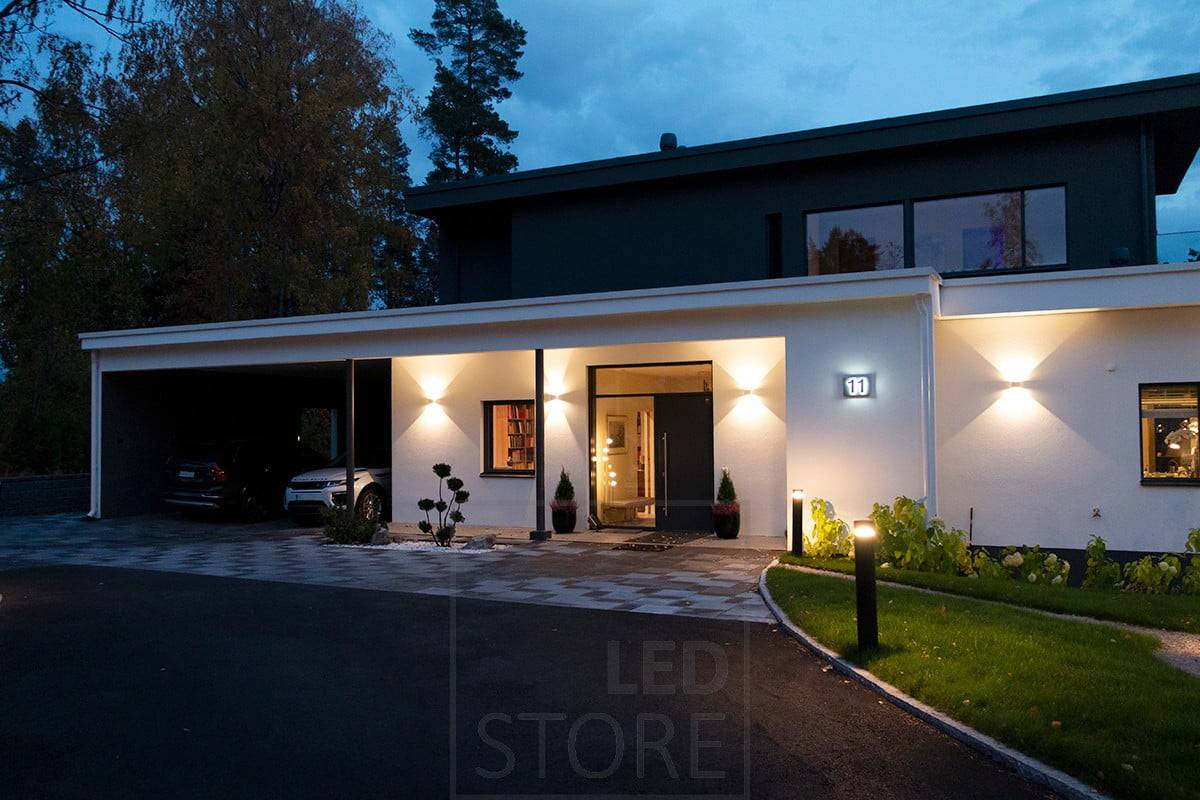 Bílé obousměrné nástěnné svítidlo FUNK osvětlující fasádu a vchod. Příjezdová cesta je osvětlena sloupovými svítidly. Ledstore.fi