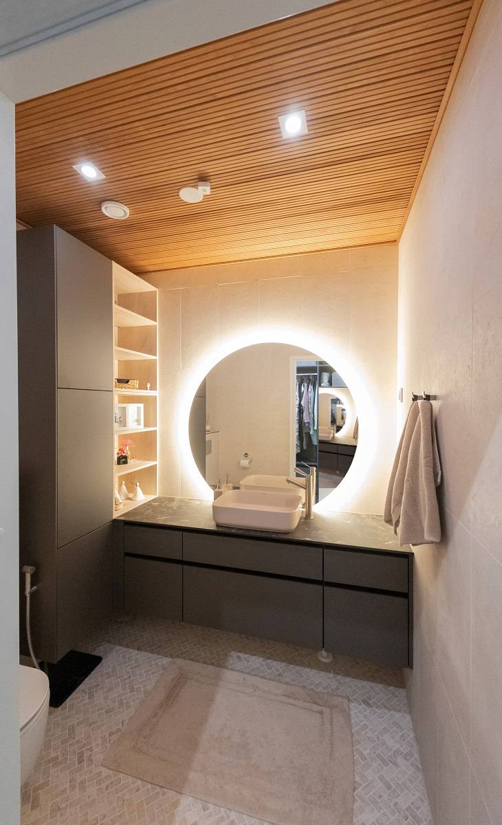 Moderní koupelna s LED osvětlením od LedStore.fi: kruhové zrcadlo s LED podsvícením nad umyvadlem vytváří elegantní atmosféru. V koupelně jsou tmavé skříňky, teplý dřevěný strop a světlé stěny. Ručníky jsou umístěny na pravé straně a vlevo na vás čekají poličky. Na podlaze spočívá béžový koberec, který umocňuje celkový dojem.