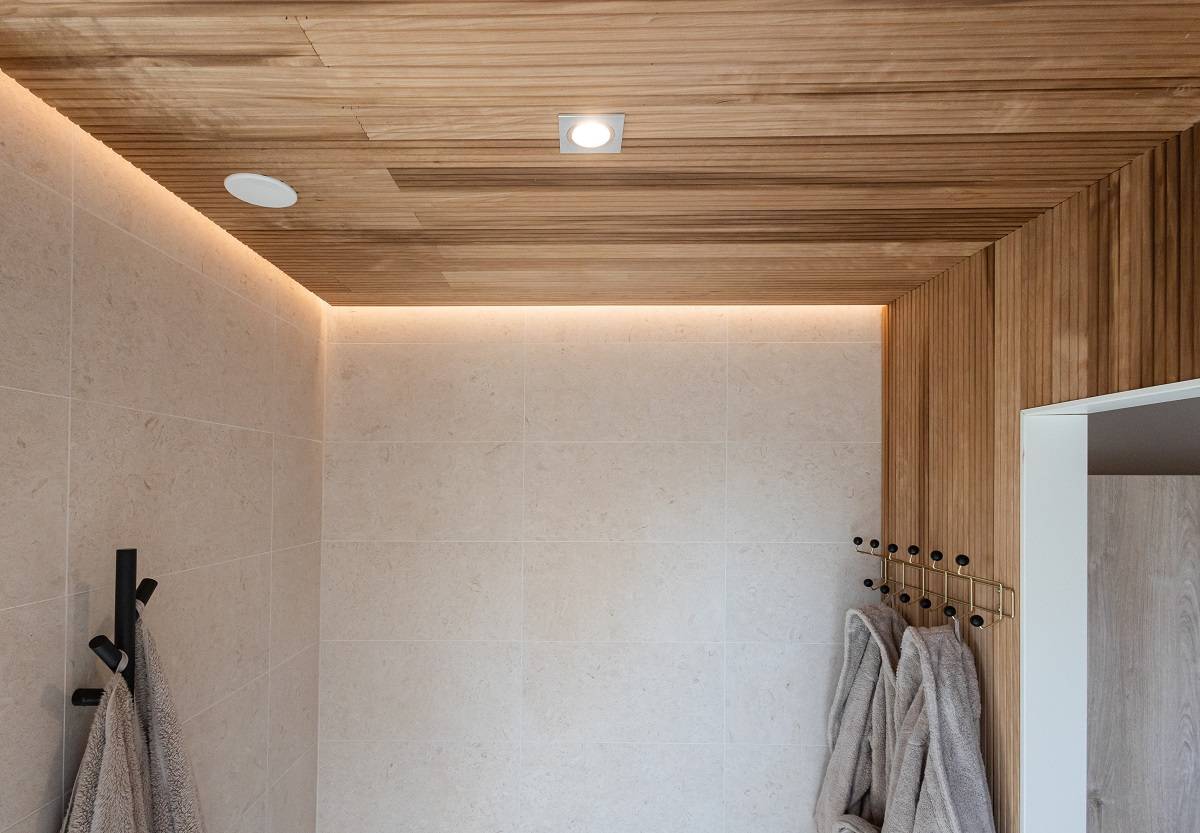 LedStore.fi nabízí řešení pro moderní koupelnu s moderním LED osvětlením. Béžové obložené stěny, dřevěný strop a zapuštěné LED osvětlení vytvářejí svěží a moderní vzhled. Měkké šedé ručníky zavěšené na černých háčcích harmonizují minimalistický interiér.