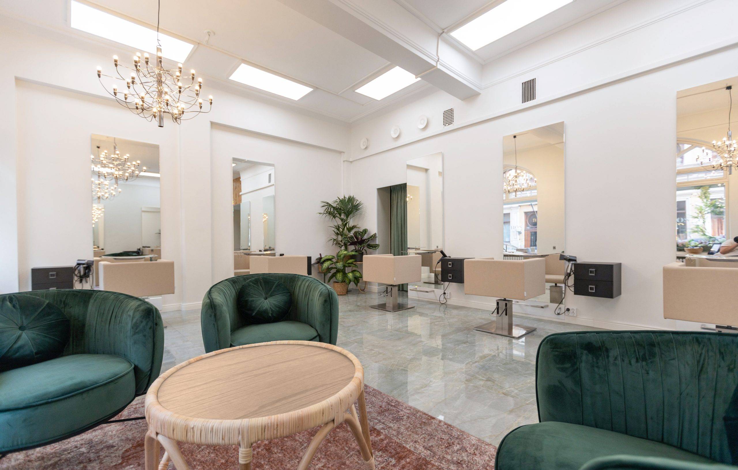 Světlý kadeřnický a holičský salon s velkými LED panely na stropě. Ze stropu visí také velký lustr. V přední části jsou křesla a malý stolek.