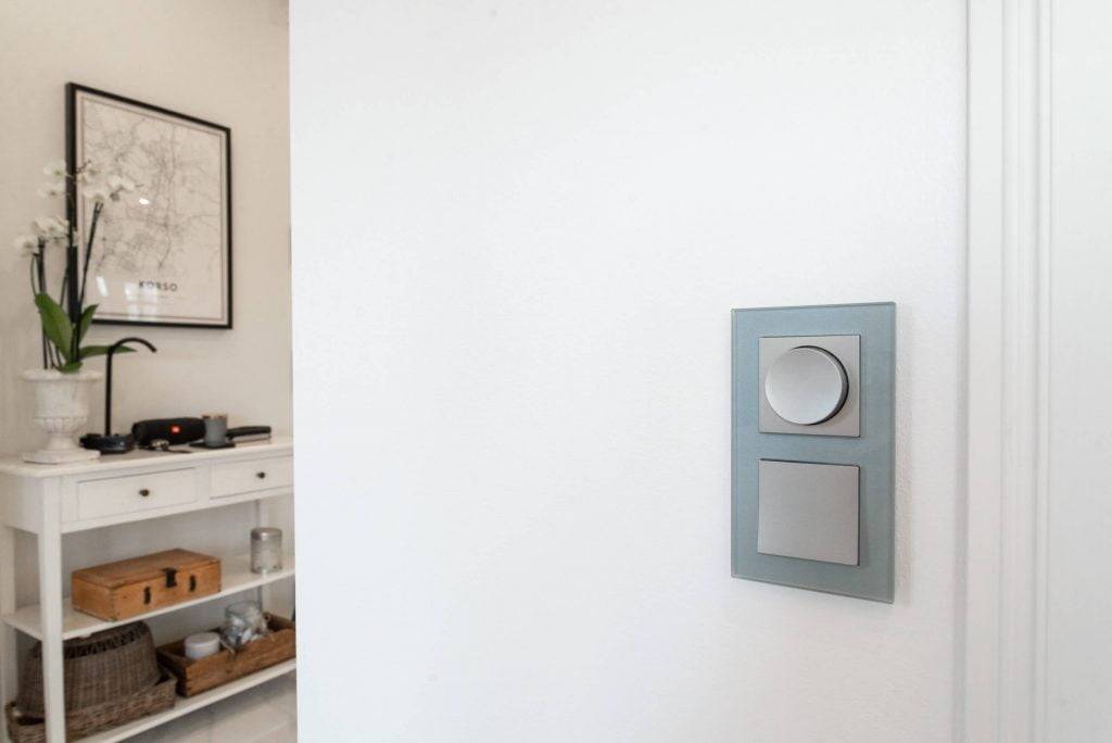 Moderní vypínač LED se dvěma tlačítky na bílé stěně; bílá konzola s orchidejí v květináči a zarámovanou mapou v minimalistickém pokoji.