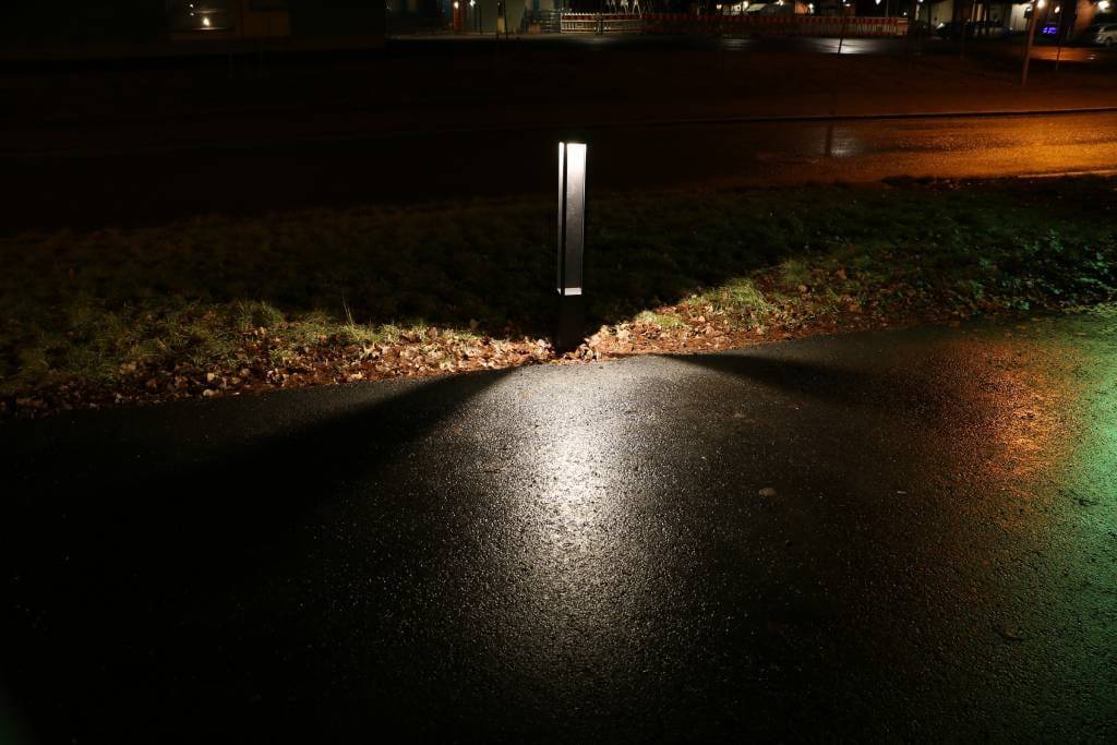 Pouliční osvětlení LED, které osvětluje mokrou ulici s trojúhelníkovými vzory ohraničenými trávou; městská kulisa se vzdálenými světly.