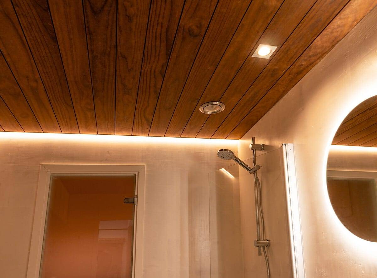 Dřevěný strop koupelny s ambientním LED osvětlením, moderní sprchovou hlavicí, bodovým světlem a kruhovým LED zrcadlem; dveře vlevo.