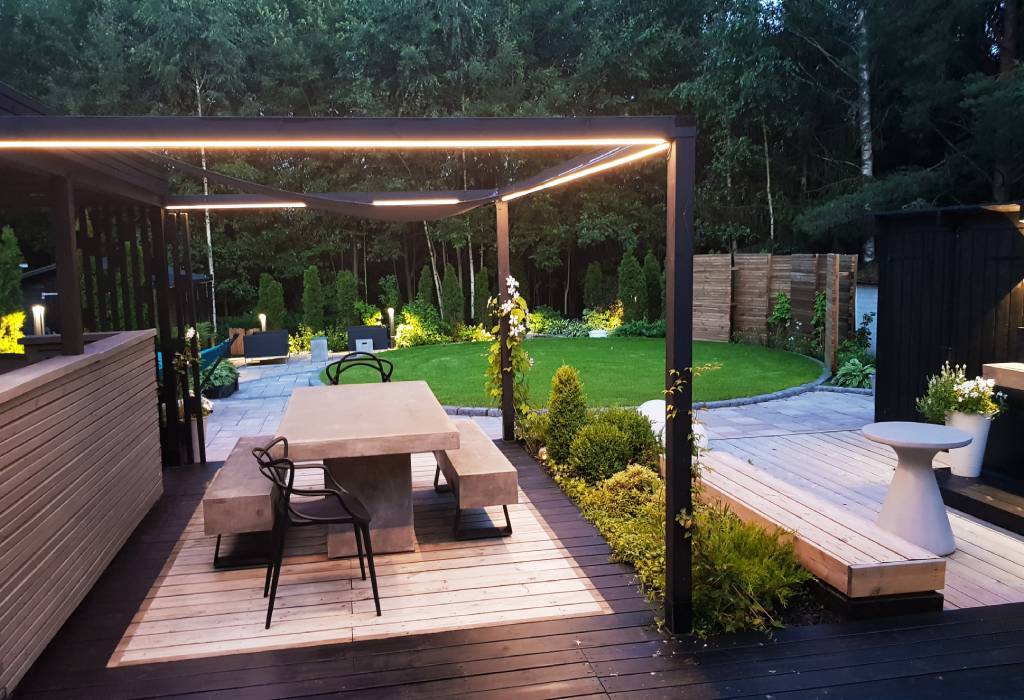 Moderní zahradní terasa s betonovým jídelním stolem a černými židlemi pod minimalistickou pergolou. Pergola má integrované LED osvětlení, které vytváří náladovou atmosféru. Plochu obklopuje zelená vegetace, jediný udržovaný trávník a kamenná cesta, což jí dodává přírodní kouzlo. Kromě toho je zde jako malý detail umístěn bílý moderní truhlík na květináče, který slouží k vychutnávání elegantní terasy. Podívejte se na širokou nabídku LED osvětlení LedStore.fi a přeneste stejnou moderní eleganci i na svůj dvůr!
