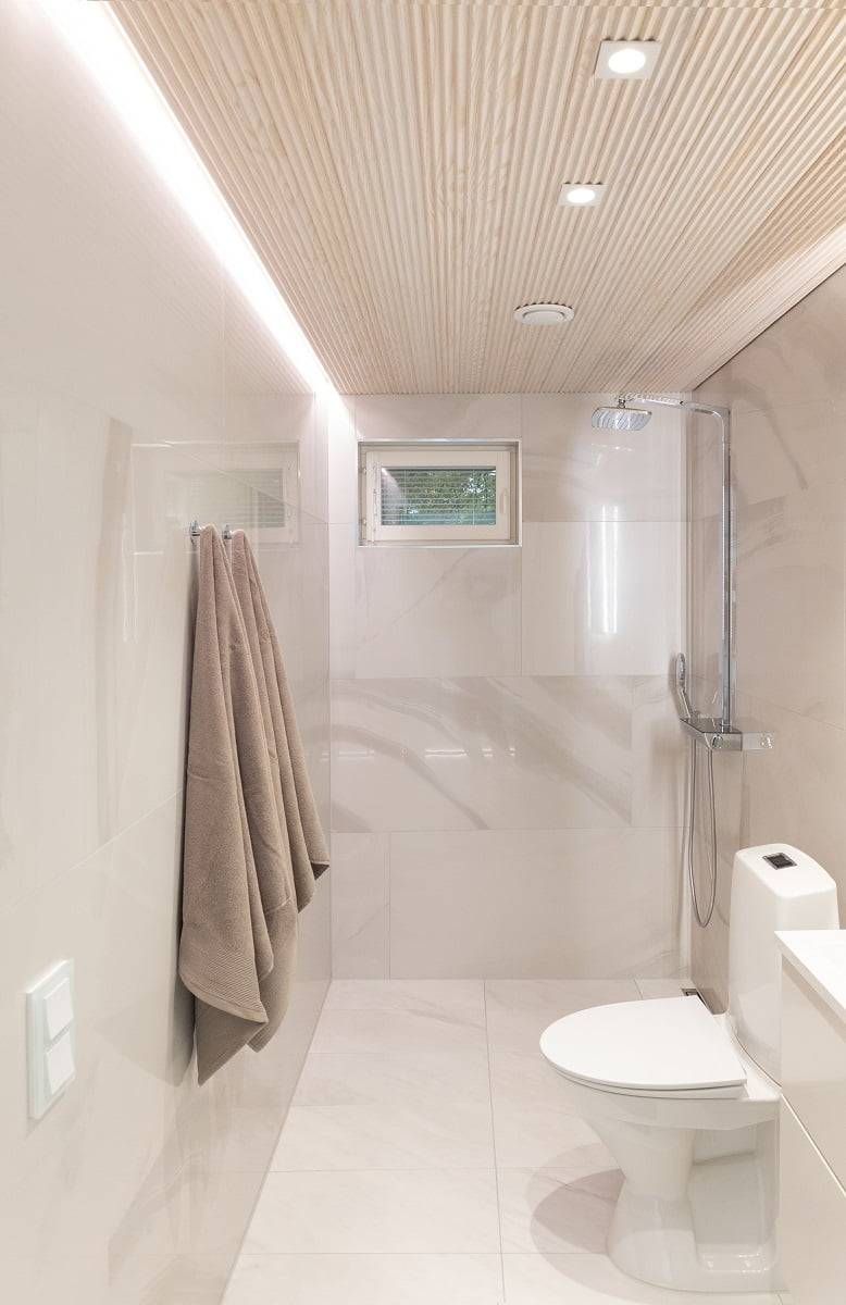 Moderní a minimalistická koupelna se světle béžovými stěnami a dřevěným obkladem. Eleganci prostoru dodává nástěnný věšák na ručníky se dvěma osuškami, malé okno a sprchový kout oddělený skleněnou přepážkou. Účinné osvětlení zajišťují integrovaná stropní LED svítidla. LedStore.fi: Objevte řešení LED osvětlení, která jsou ideální pro stylové a nadčasové interiéry.