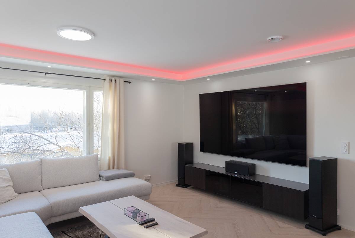 Osvětlení - Villa Vuorentausta Moderní obývací pokoj plný elegantní atmosféry. Velká pohovka vybízí k odpočinku a světlý dřevěný konferenční stolek dodává prostoru teplo. Moderní nádech místnosti dodává nástěnná televize s plochou obrazovkou a LED podsvícením. Svůj zážitek umocníte podlahovými reproduktory a vychutnáte si červené akcentní LED osvětlení na stropě. Velké okno propouští přirozené světlo, takže prostor působí prostorně a světle. Všechny tyto prvky dohromady vytvářejí dokonalé prostředí díky všestranným řešením LED osvětlení od společnosti LedStore.fi.