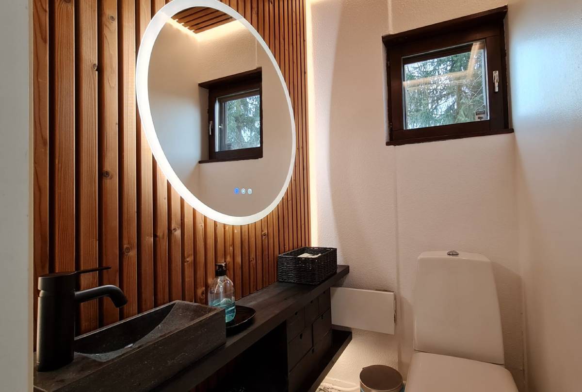 Moderní koupelna s LED osvětlením od společnosti LedStore.fi zahrnuje kruhové LED zrcadlo na dřevěné stěně. Pod zrcadlem je černá deska s obdélníkovým umyvadlem a černou baterií a dávkovačem mýdla. Nad záchodovým prkénkem je malé okénko a poblíž se nachází praktické úložné řešení.