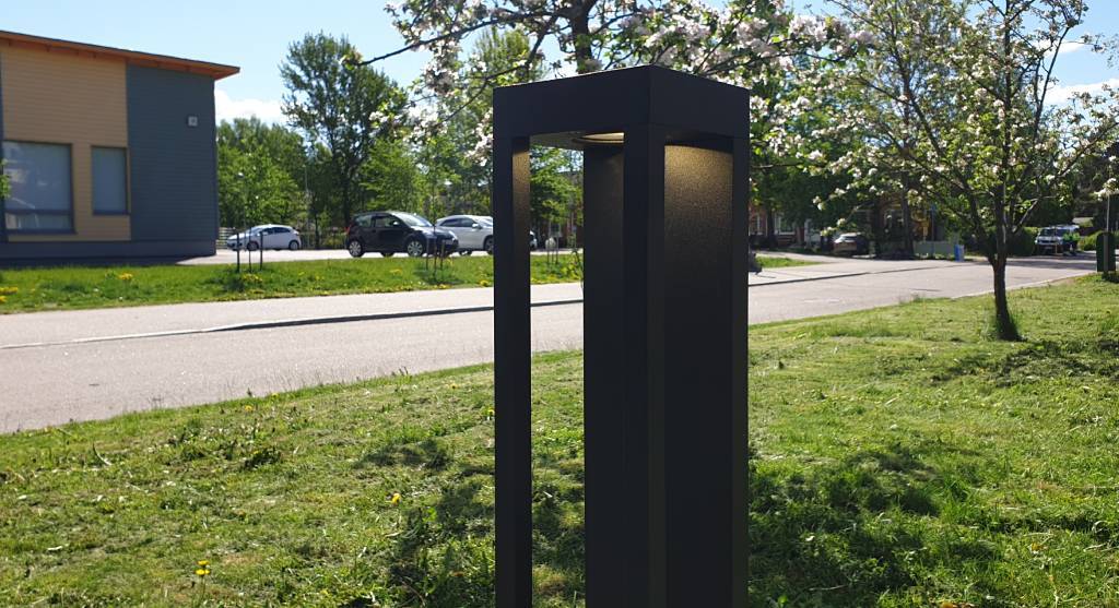 Moderní černý sloup veřejného osvětlení s LED světlem podél silnice, kvetoucí stromy, zaparkovaná auta a budova s velkými okny v pozadí pod slunečnou oblohou.