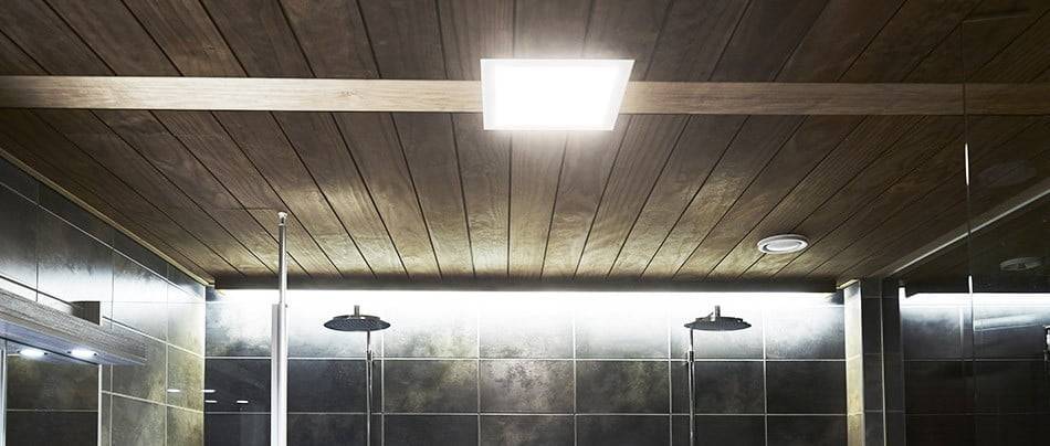 Moderní koupelna s dřevěným stropem, čtvercovým LED svítidlem, tmavými obklady, dvěma dešťovými sprchovými hlavicemi a příjemnou atmosférou s jemným LED osvětlením.