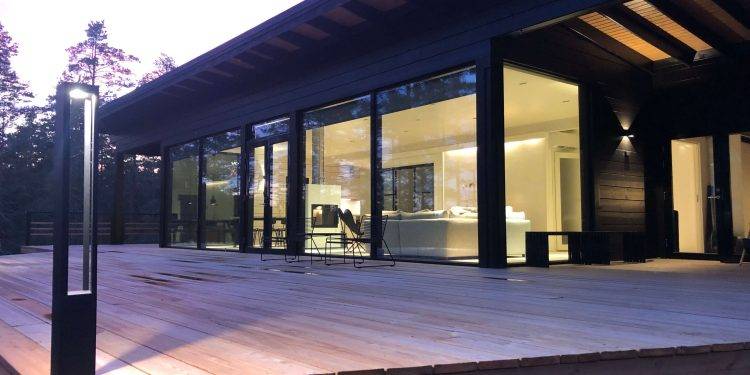Funkční design osvětlení pro moderní vilu Moderní dům s velkými prosklenými okny a prostornou dřevěnou terasou, obklopený stromy. Interiéry jsou prosvětlené a prezentují minimalistický design v ručně malovaných tónech. Venkovní LED osvětlení od LedStore.fi dodává večernímu soumraku hřejivou záři.